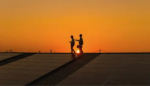 Un tejado al atardecer destaca por una extensa instalación de paneles solares bajo un cielo anaranjado. En el fondo, se vislumbra la silueta de una ciudad o un complejo industrial. La escena refleja innovación, sostenibilidad y el impacto positivo de la energía solar fotovoltaica en el entorno empresarial e industrial.